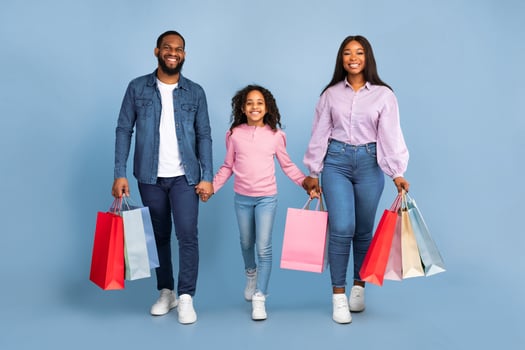 Family Shopping Time. Happy black man, woman and their daughter walking together with colorful shopping bags, holding hands and looking at camera isolated over blue background in studio photo