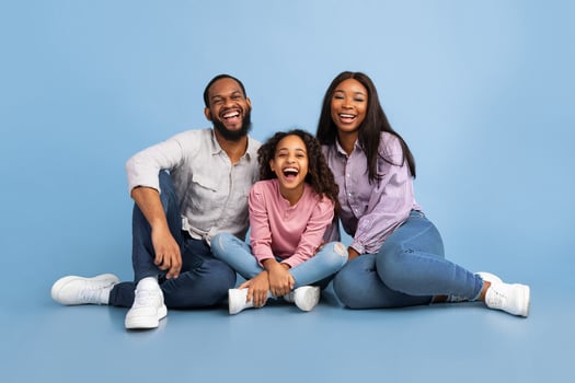 Portrait of excited black man, woman and little girl sitting on the floor isolated over blue studio wall. Cheerful daughter, mother and father laughing and looking at camera. Bonding Concept photo