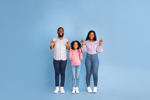 Cheerful African American Family Of Three People Standing On Blue Background. Excited Man, Woman And Adolescent Girl Pointing Finger Up Showing Free Copy Space. Empty Place For Advert, Full Length photo