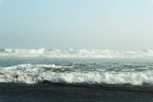 A serene coastal scene with powerful waves crashing onto a dark, sandy shore. The vast expanse of the ocean meets a clear, blue sky, creating a breathtaking panorama photo