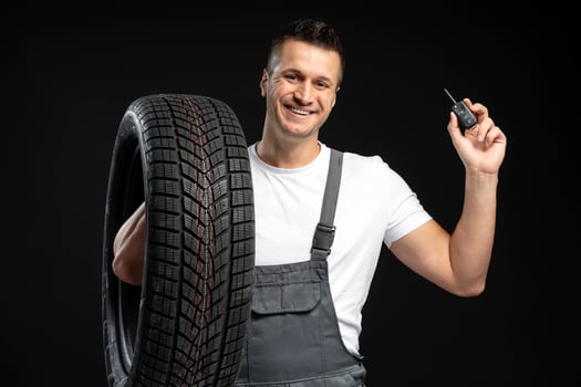Mechanic holding car tire and key remote, smiling on black background photo