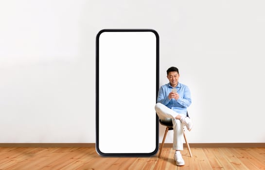 Asian man in a light blue shirt and white sneakers relaxes in a chair beside a large blank smartphone screen, set in a contemporary room with wooden floors and a minimalistic design. photo