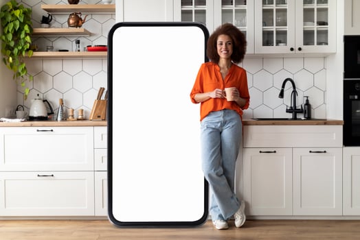 African American woman in an orange blouse leans against a large blank screen while holding a cup of coffee in a stylish kitchen filled with natural light and contemporary decor. photo