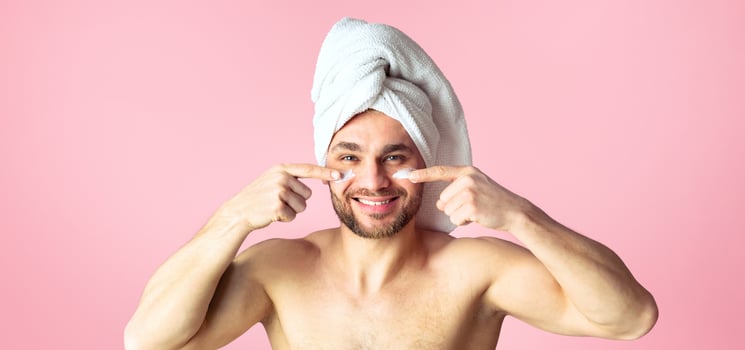 Male body care concept. Cute young man after a shower, white towel on head, smears cream on face. photo