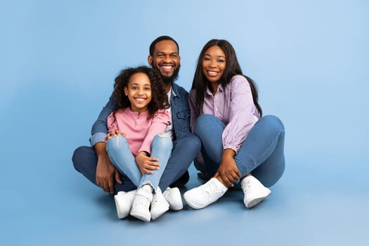 Happy Loving Family. Portrait of African American man, woman and girl sitting on the floor isolated on blue studio wall. Smiling couple posing with daughter looking at camera. Togetherness And Bonding photo