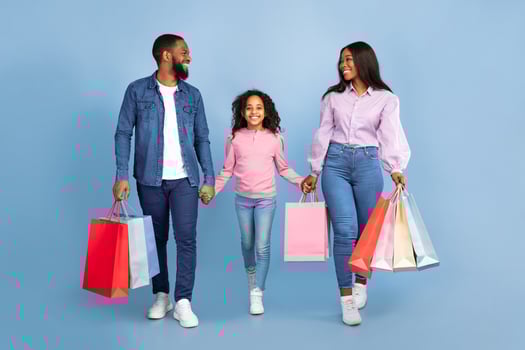 Happy black man, woman and their cute little daughter walking together with colorful shopping bags, holding hands and looking at each other isolated over blue background in studio, copy space photo