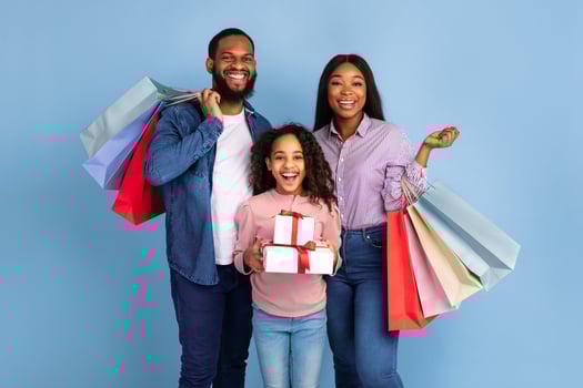Portrait of smiling African American family of three people carrying colorful shopping bags, cheerful curly girl holding stack pile of gift boxes with present, isolated over blue studio wall photo