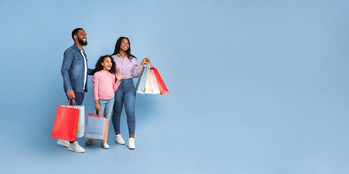 Family Shopping. Full body length portrait of African American cheerful people holding shopping bags with new clothes, excited girl, guy and lady looking aside at free copy space, blue studio panorama photo