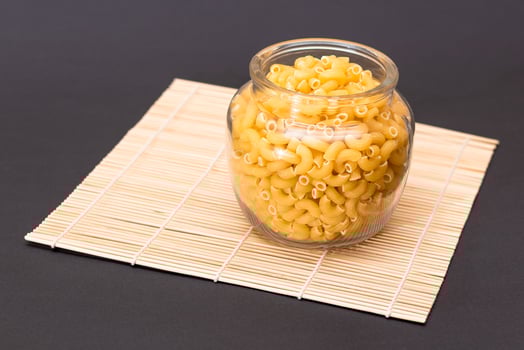 Uncooked Chifferi Rigati Pasta in Glass Jar on Bamboo Mat on Black Background. Fat and Unhealthy Food. Classic Dry Macaroni. Italian Culture and Cuisine. Raw Pasta photo