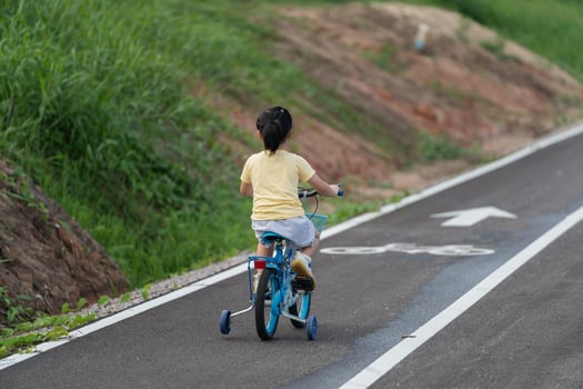 Asian young girl is riding a blue bicycle down a road. The road is lined with grass and dirt, and there is a white arrow on the side of the road. The girl is wearing a yellow shirt photo