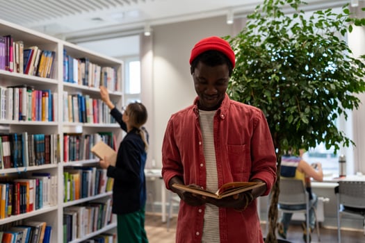 Smiling student black guy stands in library read book in background girl reach for top of bookshelf. photo