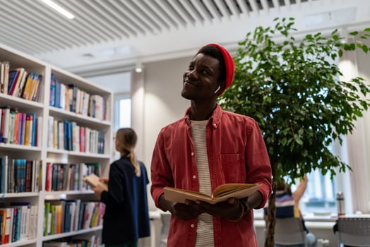 Joyful black student guy stands in university library with book thinks about good job after diploma. photo