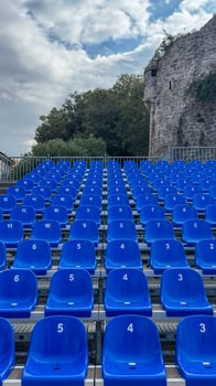 Rows of blue plastic numbered seats in an outdoor seating area, backed by trees and rock formations photo