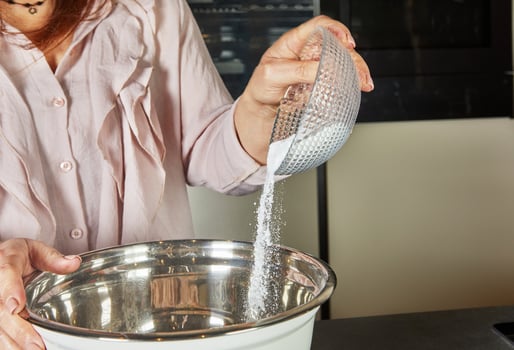 A person pours granulated sugar from a small glass bowl into a large metal mixing bowl, adding sweetness to the recipe. photo