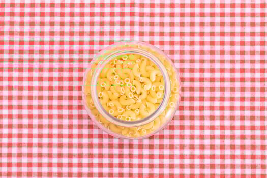 Uncooked Chifferi Rigati Pasta in Glass Jar on Checkered Red Towel on Pink Background. Fat and Unhealthy Food. Scattered Classic Dry Macaroni. Italian Culture and Cuisine. Raw Pasta photo