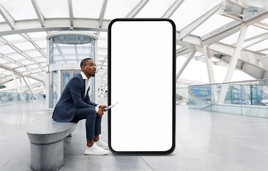 African American man in a suit is seated on a bench in a contemporary architectural space, appearing thoughtful as he waits next to a large, empty digital display. photo