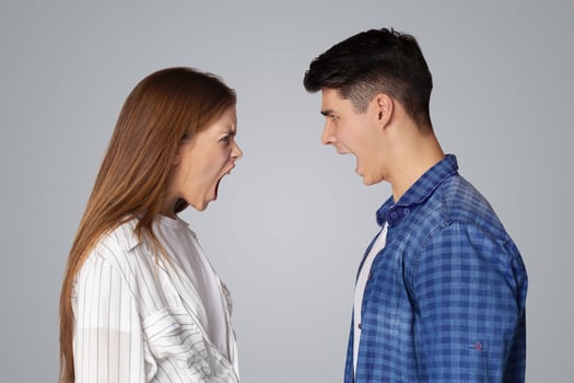 Breakup of relations, quarrel, problems in couple and scandal. Angry aggressive young man and lady yelling at each other with open mouths, isolated on gray background, studio shot, profile, free space photo