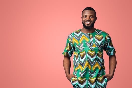 Handsome black man in traditional african clothes smiling over blue studio background, putting hands in pockets, copy space. Cheerful african american guy posing in ethnic colorful clothes photo