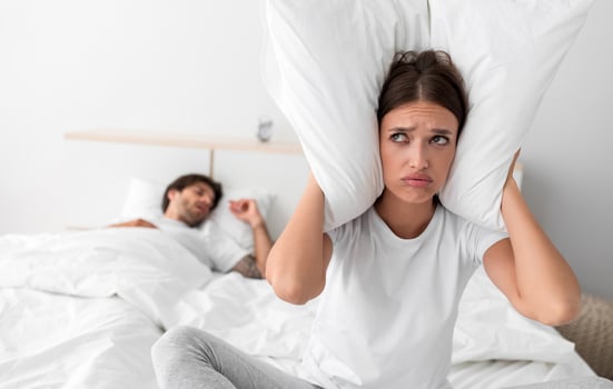 Despaired millennial european wife sits on bed covers ears with pillow and suffers from snoring of sleeping husband in bedroom interior. Intolerable snoring and noise at night, insomnia and apnea photo