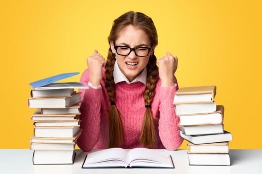 Heavy study, many tasks and lessons. Tense teenager girl in glasses with stacks of books on table isolated on blue background, studio shot photo