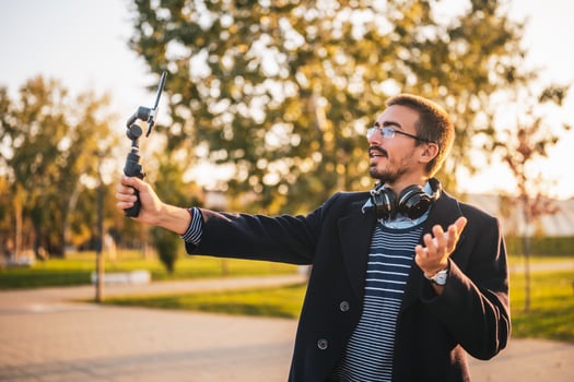 Businessman vlogger in coat wearing eyeglasses and headphones. He is using mobile phone with gimbal in the park and talking while recording vlog on a sunny autumn afternoon. photo