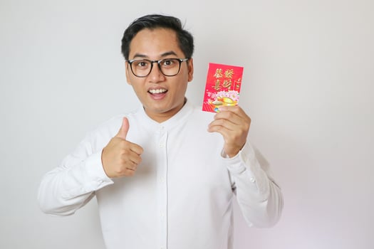 Smiling young Asian men wearing a traditional cheongsam qipao dress holding angpao or a red packet of monetary gift and showing thumbs up gesture isolated on white background. Happy Chinese New Year. photo