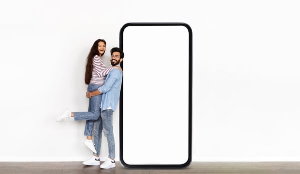 Indian couple stands next to an oversized blank phone display in a well-lit indoor setting, with one partner playfully lifted off the ground, showcasing a fun and dynamic moment. photo