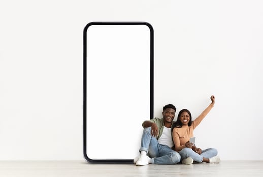 A cheerful black couple sits on the floor, relaxed and smiling. They lean against a large blank phone screen, enjoying their time together and engaging in conversation. photo