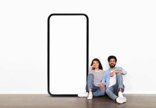 A cheerful young Indian couple relaxes together against a clean white wall, sitting next to an oversized smartphone frame that is ready for creative displays or promotions. photo