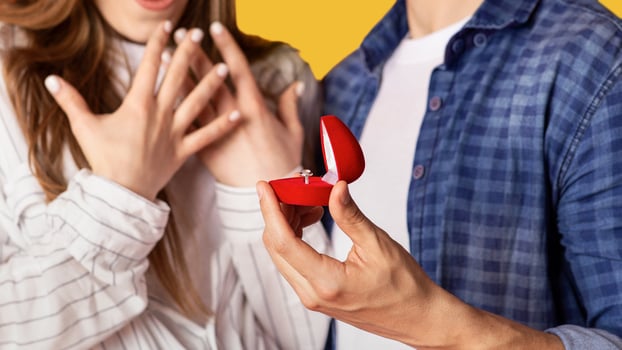 Marriage proposal and people emotions. Focus on box with ring in strong man hand and excited gestures of happy and surprised woman, isolated on yellow background, studio shot, close up, cropped photo