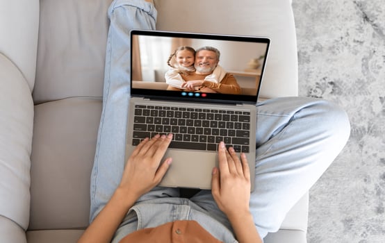 A person sits comfortably on a couch using a laptop for a call with a smiling relative. The warm atmosphere suggests a family gathering despite the distance. photo