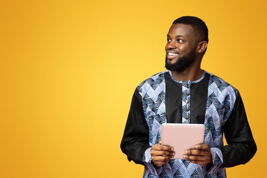 Smiling african american guy using modern digital tablet, wearing african t-shirt, pink studio background. Young black man in traditional costume holding pad and looking at copy space, panorama photo