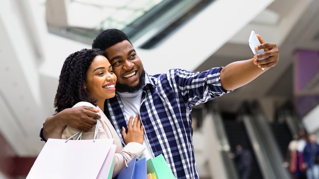 Shopping Fun. Joyful black couple taking selfie in mall center, using smartphone, holding lots of paper bags photo