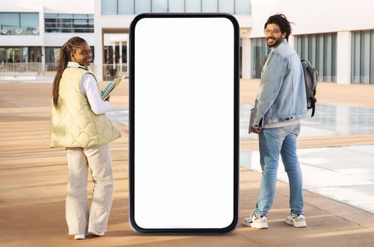 Two young adults pose outdoors beside a large blank screen, enjoying a sunny day. They are fashionably dressed, with one holding a notebook while smiling at the camera. photo