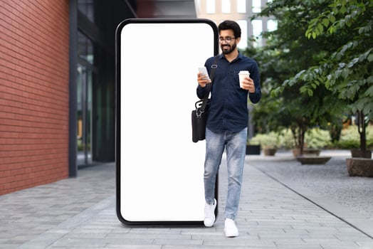 A young Indian man in a casual outfit holds two coffee cups while walking past a tall blank digital screen in a modern city environment. The scene captures a blend of technology and urban life. photo