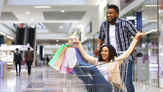 Department Store Fun. Happy african couple enjoying shopping together, man pushing shopping cart with girlfriend inside, fooling in mall, free space photo