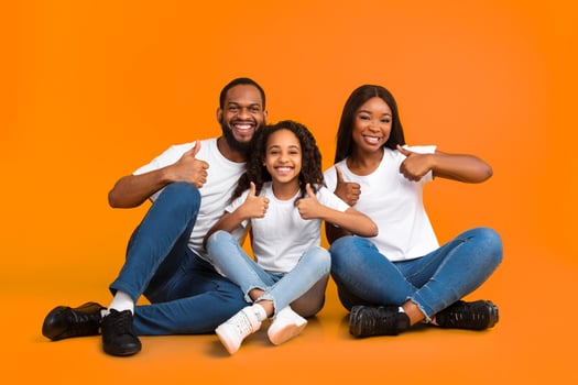 Family Approval. Portrait of smiling black family showing thumbs up sign gesture with both hands, recommending something cool. Mom, dad and daughter sitting on the floor at yellow studio photo