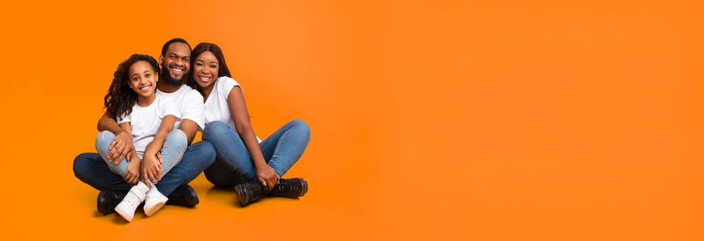 Loving Family. Casual African American man, woman and girl sitting on floor isolated on yellow orange wall. Smiling couple posing with daughter looking at camera, panorama, banner, free copy space photo