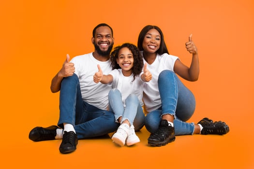 We Approve And Recommend. Excited Black Man, Woman And Teen Girl Gesturing Thumbs Up Okay Looking At Camera Sitting On The Floor Over Yellow Orange Studio Background. Avertisement And Support Concept photo