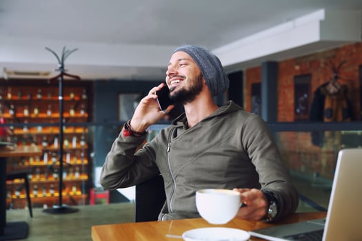 Young handsome hipster man with beard sitting in cafe talking mobile phone, holding cup of coffee and smiling. Laptop on wooden table. photo