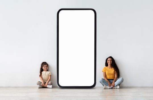A woman and a young girl are seated on a smooth floor next to a large blank smartphone display against a light-colored wall. They appear relaxed and engaged with each other. photo