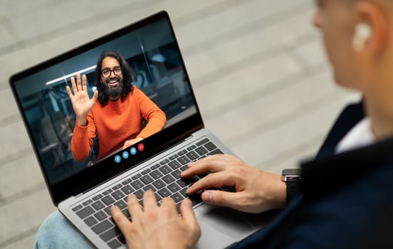 A person sits at a laptop, participating in a call with someone wearing an orange sweater. The environment suggests a modern workspace, enhancing the social interaction. photo