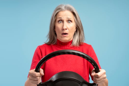 Sad, fear senior woman holding steering wheel looking at camera standing isolated on blue background photo