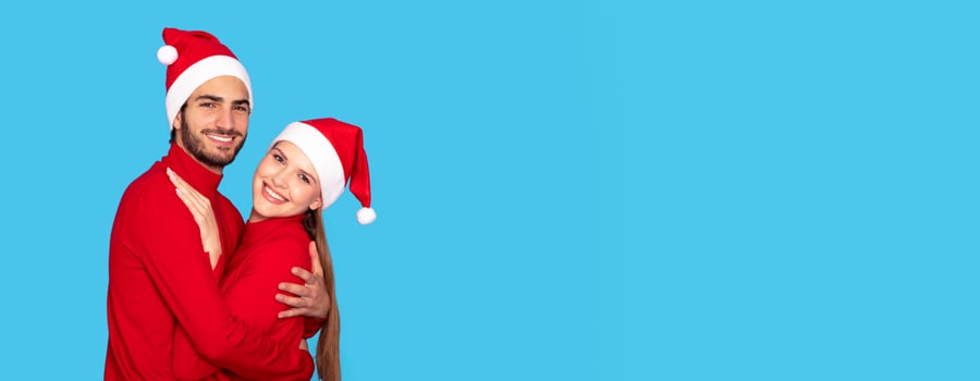 Hugging Young Couple In Santa Hats Posing Over Blue Background In Studio, Cheerful Happy Millennial Man And Woman Embracing And Smiling At Camera, Spending Christmas Holidays Together, Copy Space photo
