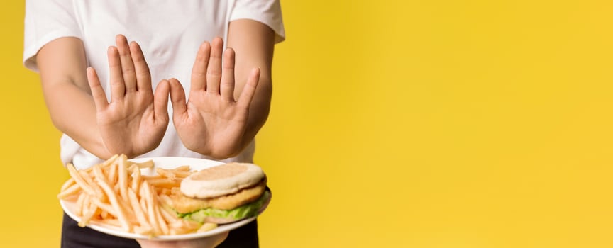Unrecognizable Woman Rejecting Consume Plate With Junk Food, Showing Stop Gesture With Open Palms, Yellow Background, Cropped Image photo