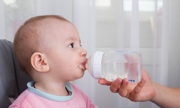 A mother's hand gives water to a baby to drink from a bottle while feeding porridge. A child boy aged one year drinks water. Copy space for text, caucasian photo