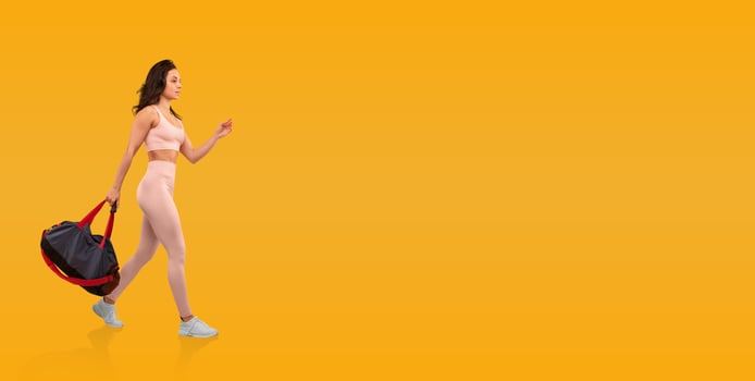A young woman confidently strides forward, carrying a large duffel bag while wearing minimal activewear. The bright yellow background enhances her dynamic movement and fitness vibe. photo