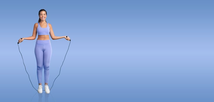 A young woman is happily skipping rope, wearing comfortable workout attire in a solid blue background. She appears engaged in a fitness routine, promoting an active lifestyle and health. photo