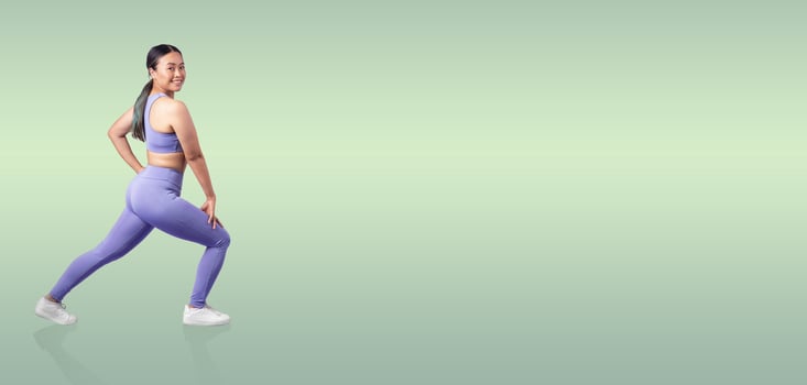 A fit woman performs a lunging exercise while wearing stylish activewear. She smiles confidently against a soft, soothing green background, promoting fitness and health. photo