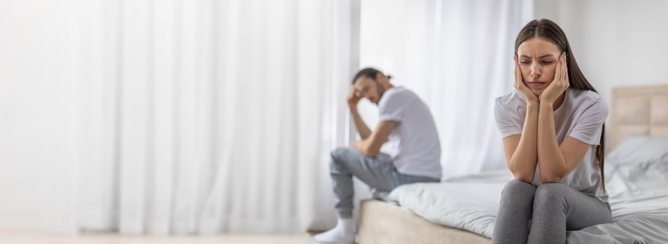 A couple sits separately on a bed, visibly distressed. The woman holds her head in her hands while the man appears frustrated, as they experience a difficult conversation in their bedroom. photo